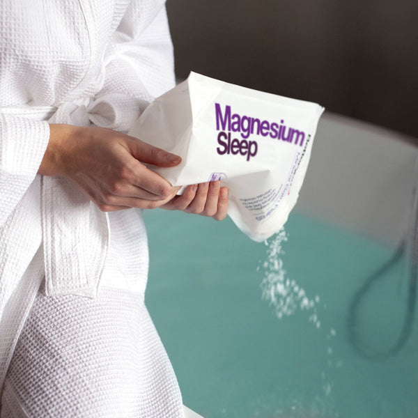 A woman in a bath robe pouring magnesium sleep flakes into a full bath tub