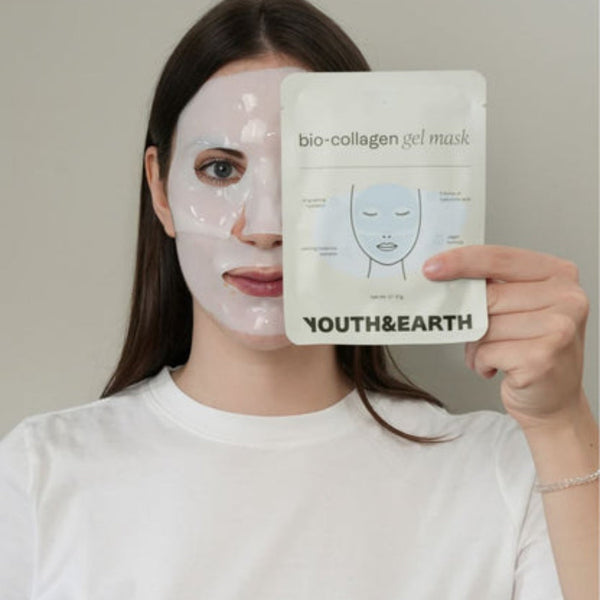 A woman wearing a white face mask holds up a sheet of Youth & Earth bio-collagen gel mask in front of her face against a plain light gray background.