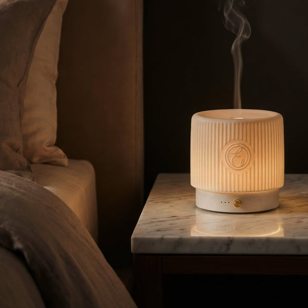 White ceramic diffuser emitting steam on a marble nightstand next to a bed.