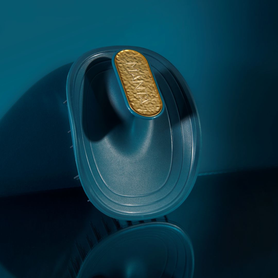 A close-up studio shot of a dark teal hairbrush with a textured gold oval detail on the handle, set against a matching dark teal background.