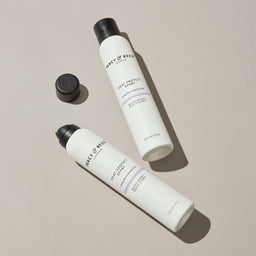 Two white bottles of FRACT OF REEFS hair protectant spray with black caps, one bottle shown with its cap off on a neutral beige background.