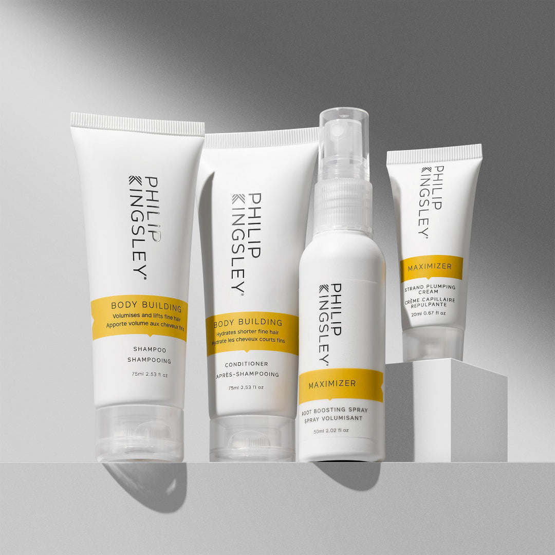 A set of four Philip Kingsley Body Building haircare products arranged on a white pedestal against a gray gradient background. The products include shampoo, conditioner, hot scoring spray, and maximizer in white squeeze bottles and tubes with yellow bands and black text.
