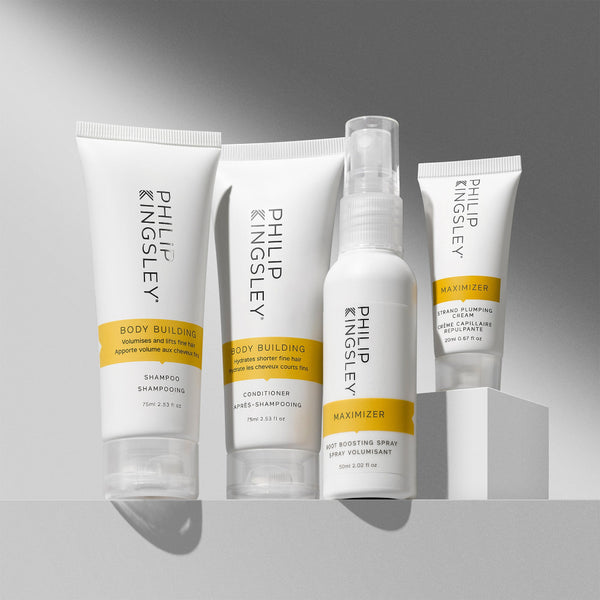 A set of four Philip Kingsley Body Building haircare products arranged on a white pedestal against a gray gradient background. The products include shampoo, conditioner, hot scoring spray, and maximizer in white squeeze bottles and tubes with yellow bands and black text.