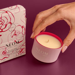 Neom scented candle with a hand holding it and the packaging on a purple background