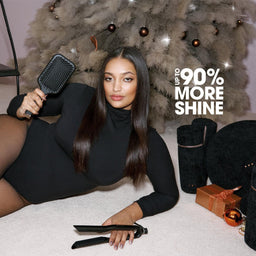 A woman in a black bodysuit and sheer tights reclines on a light carpet, holding a black hairbrush and hair straightener. Several black cylindrical hair accessories are arranged beside her, with a large fluffy gray decorative object behind her and a text overlay reading 'UP TO 90% MORE SHINE'.