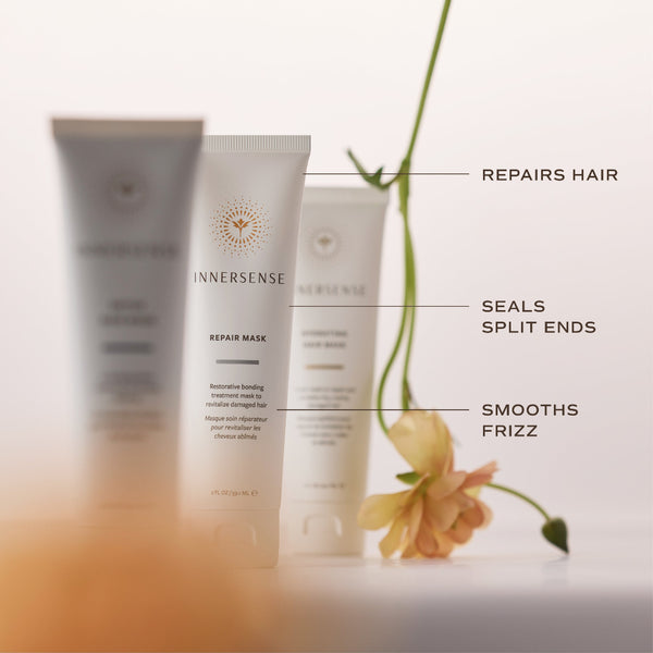 Three Innersense Repair Mask hair care tubes arranged on a white surface with decorative text callouts reading 'REPAIRS HAIR', 'SEALS SPLIT ENDS', and 'SMOOTHS FRIZZ', plus a soft peach flower prop.