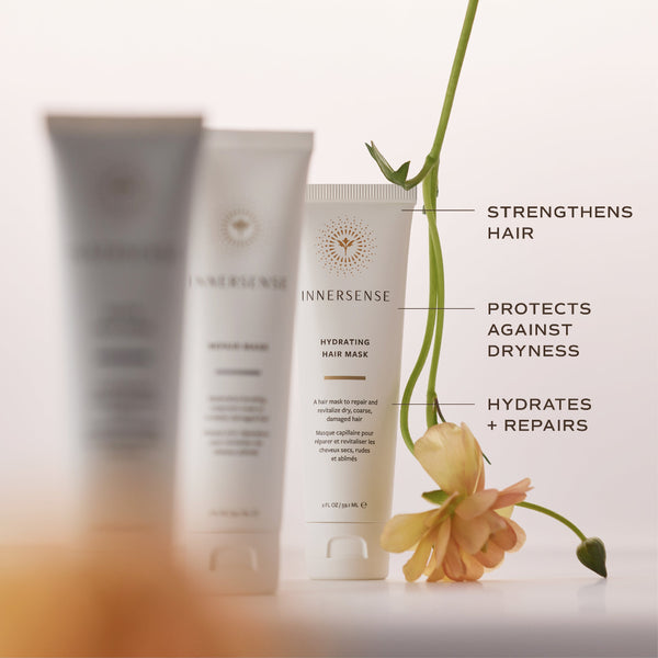Three Innersense hair care tubes on a white surface with decorative text callouts and a peach flower. The center tube is in focus and reads 'Hydrating Hair Mask,' while the two tubes behind it are blurred.