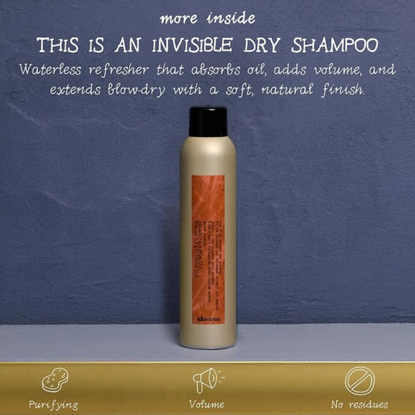 A single dry shampoo bottle stands centered against a textured blue-gray background with promotional text above and a gold strip at the bottom. The bottle is metallic champagne beige with a black cap and an orange label.