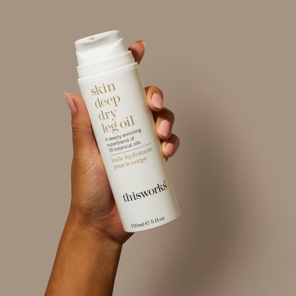 Hand holding a bottle of 'this works' skin deep dry leg oil against a beige background