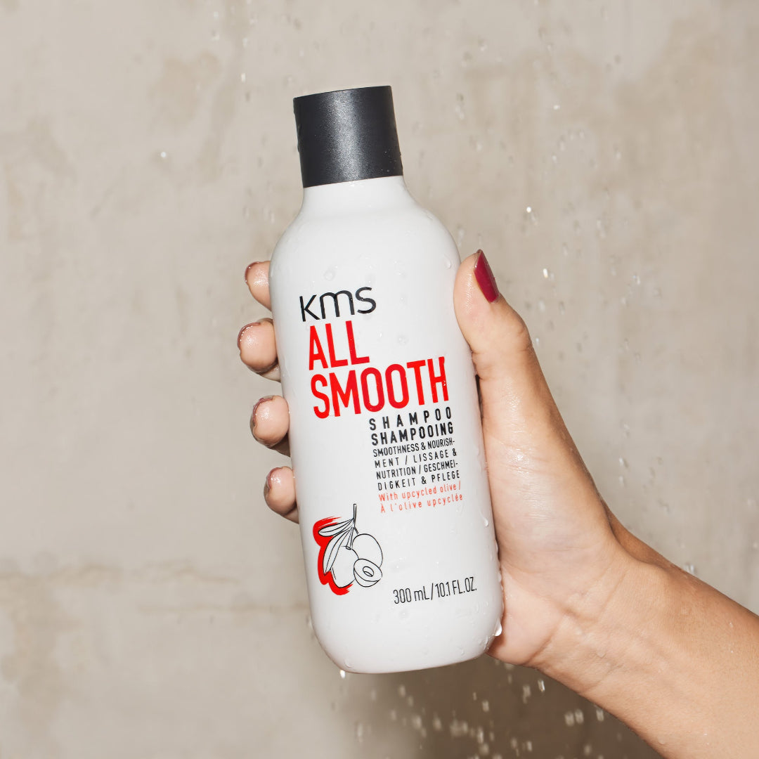 Hand holding a bottle of KMS All Smooth shampoo against a neutral background