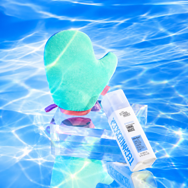 A teal and pink silicone pool float with a built-in bottle holder, shown floating on blue water with sunlight ripples. A white TechnoCheekers sunscreen bottle is resting inside the float.