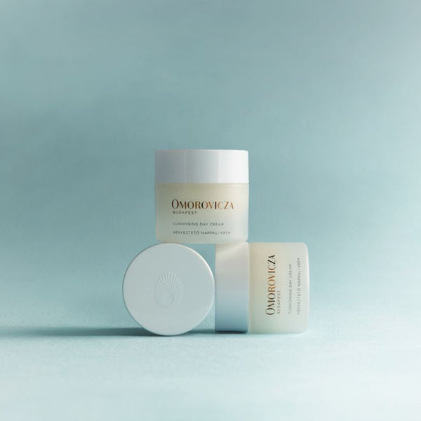 Three Omorovicza skincare containers arranged on a soft light blue studio background: a tall frosted jar, a small rectangular box, and a round compact lid in front.