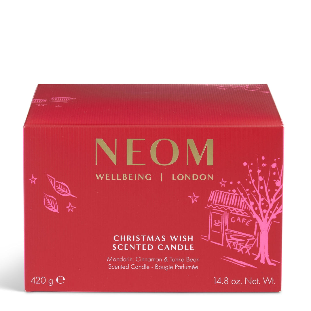 A red rectangular box of NEOM Wellbeing London Christmas Wish Scented Candle, featuring gold and pink lettering and illustrations of a tree and cafe on a white background.