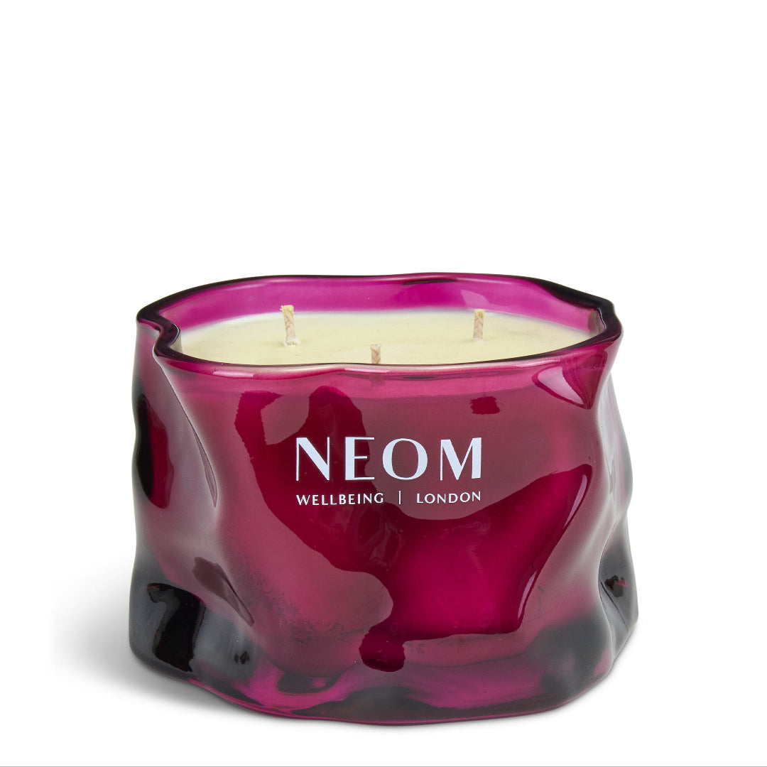 A three-wick scented candle in a wavy, organic-shaped translucent glass jar. The jar has a deep magenta-purple color with a glossy finish and the brand name 'NEOM WELLBEING | LONDON' printed in white on the front.