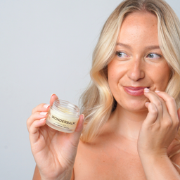 Woman holding a jar of 'Wonderbalm' and applying it to her face against a plain background