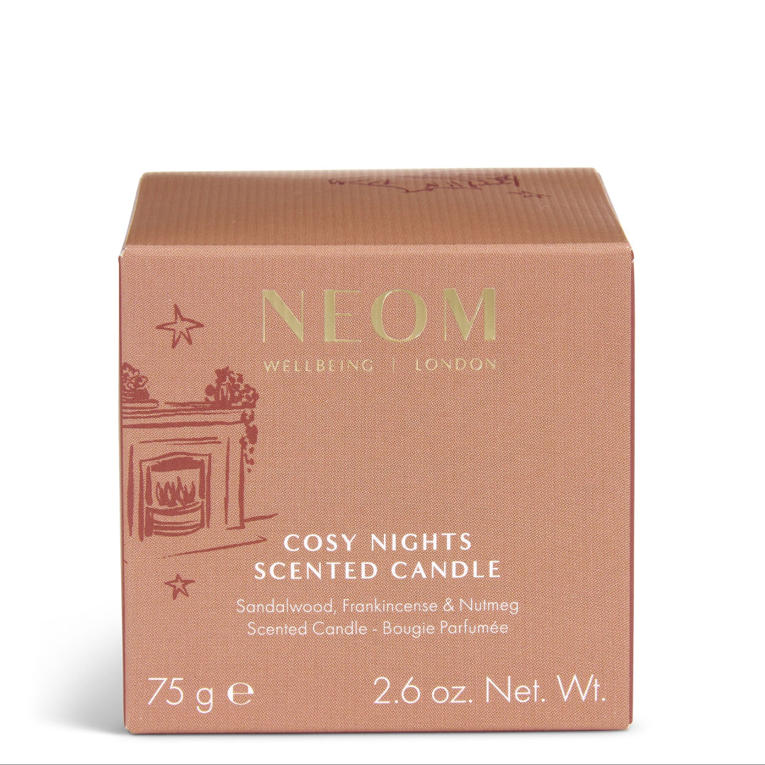 A square cardboard scented candle box by NEOM, labeled 'Cosy Nights Scented Candle,' in a dusty rose color with gold lettering and a small fireplace illustration on the front, photographed on a clean white background.