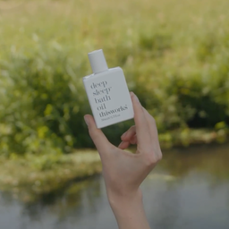 A hand holds a white rectangular bottle of thisworks Deep Sleep Bath Oil against a soft, blurred outdoor background of green grass and water.