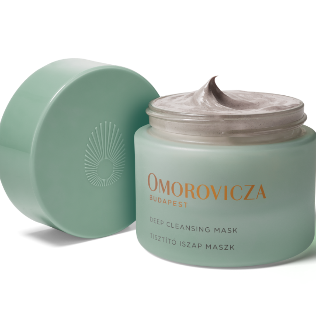 A jar of Omorovicza Deep Cleansing Mask with its lid off, showing the pale mint green jar filled with a thick, light-colored cream, and a matching lid leaning against it on a white background.