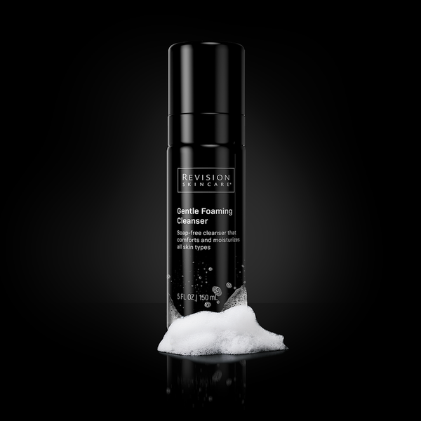 A black bottle of Revision Skincare Gentle Foaming Cleanser standing upright on a glossy black surface with white foam piled at its base.