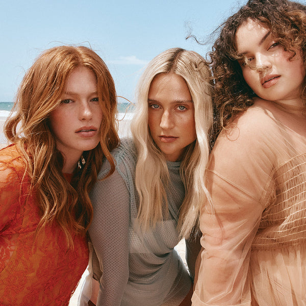 Three women standing together on a beach with clear blue sky.
