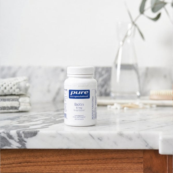 A white bottle of Pure Encapsulations Biotin 8 mg dietary supplement, set on a white marble countertop with a blurred bathroom background.