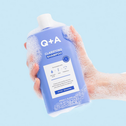 Hand holding a bottle of Q+A Clarifying Shampoo against a light blue background