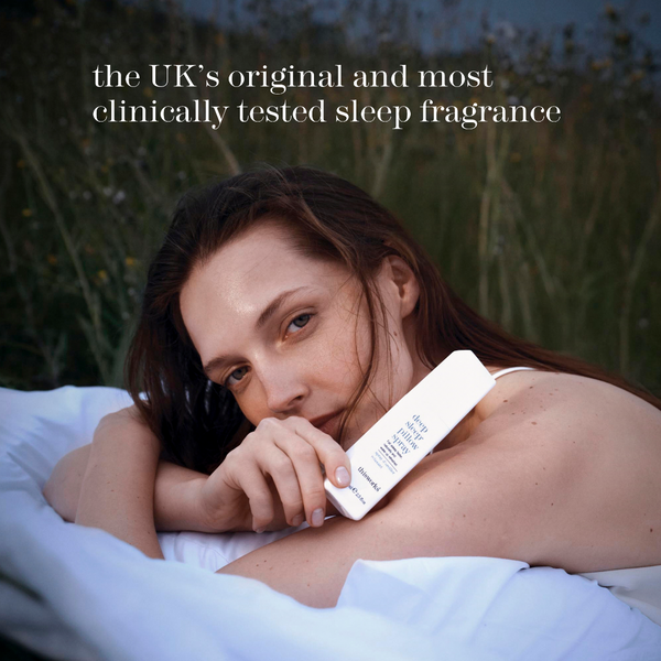 A woman lying on a white bed outdoors at dusk, holding a white rectangular sleep fragrance diffuser or perfume bottle against her shoulder. Tall grasses and a dark blue sky are blurred in the background, with white text at the top reading, 'the UK's original and most clinically tested sleep fragrance'.
