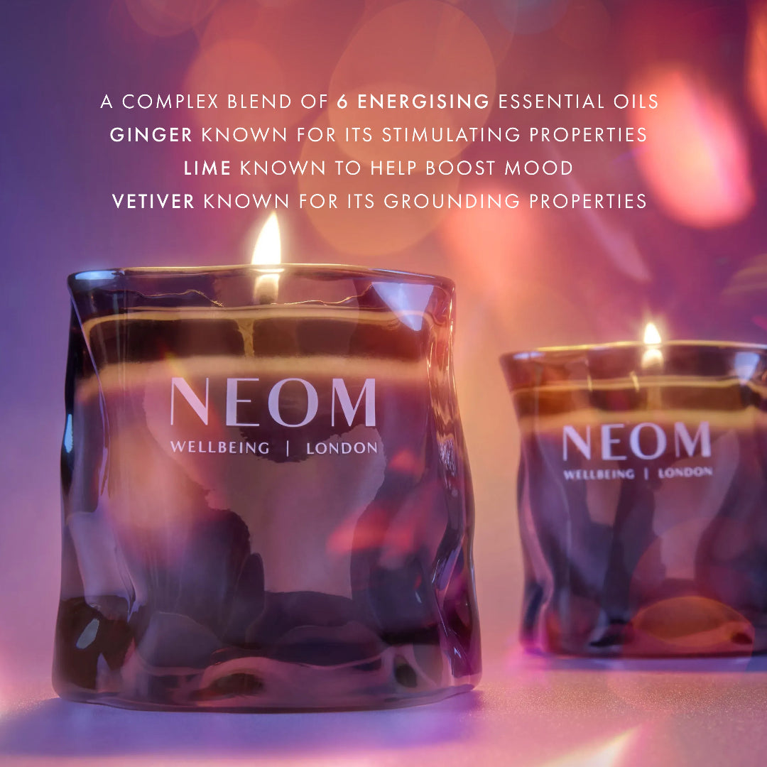 Two NEOM Wellbeing candles in textured glass holders, one in the foreground with a lit flame and one blurred in the background, set against a warm purple and orange bokeh background with descriptive text at the top.