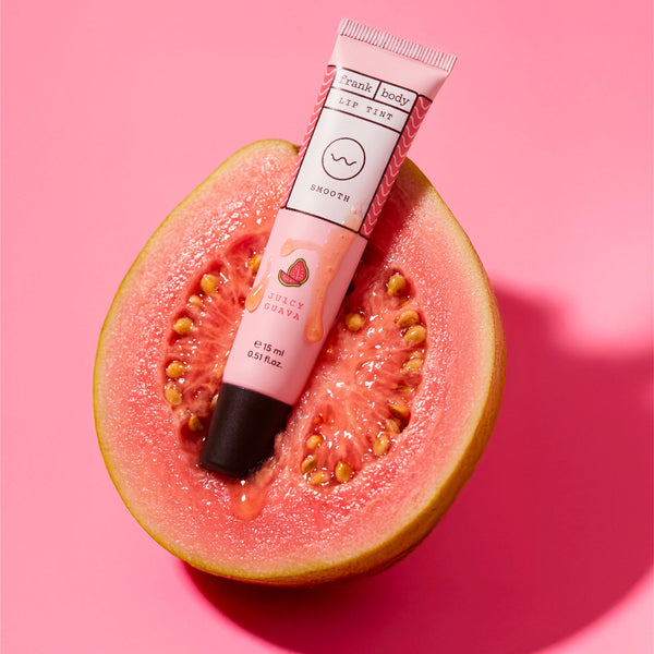 A pink and white tube of Frank Body lip tint, labeled 'Guava' and 'Sweet Guava', is placed diagonally on top of a sliced watermelon half against a solid pink background.