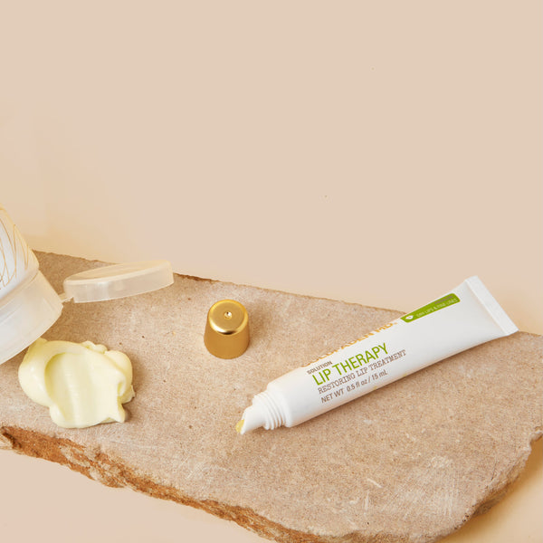 Two Golden Realm skincare tubes, a squeeze of pale yellow cream, and a gold cap arranged on a textured stone slab against a warm beige background.