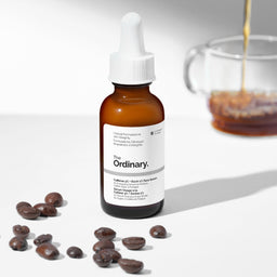 The Ordinary Caffeine + Beadie + Face Serum in an amber glass dropper bottle, styled with scattered coffee beans and a blurred glass mug of coffee on a white background.