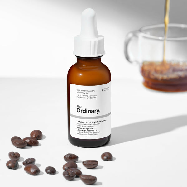 The Ordinary Caffeine + Beadie + Face Serum in an amber glass dropper bottle, styled with scattered coffee beans and a blurred glass mug of coffee on a white background.