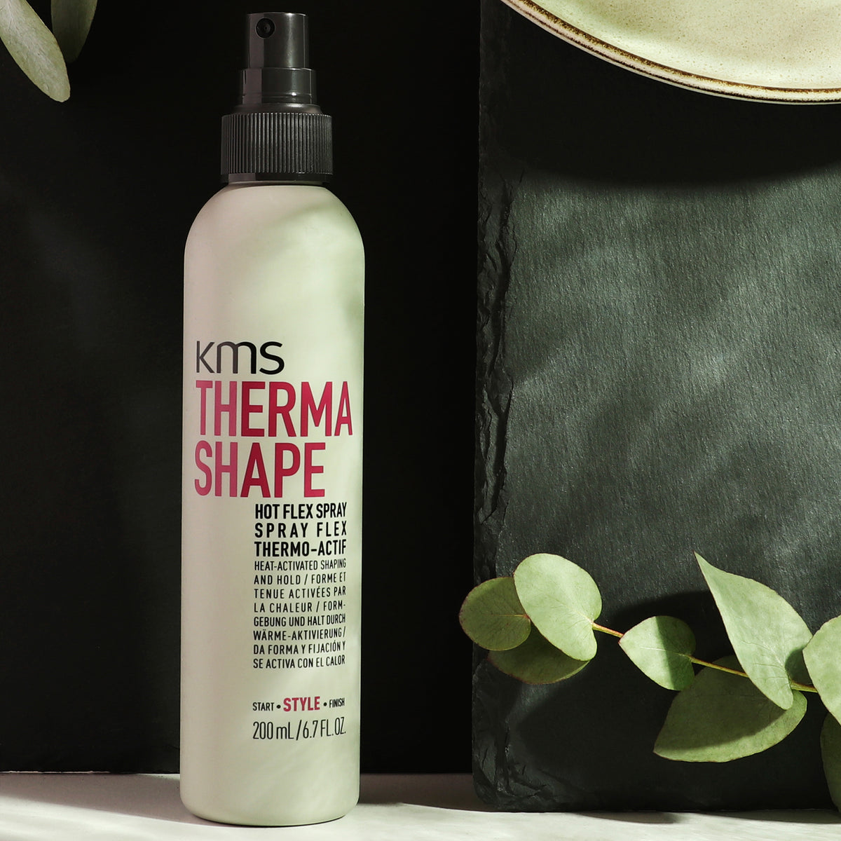 KMS Thermo Shape hair spray bottle with green leaves on a dark background