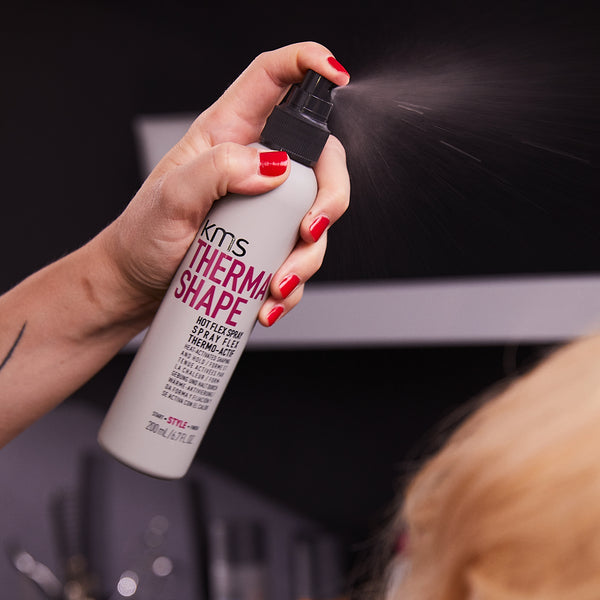 Person using a KMS Thermal Shape product on hair with a blurred background