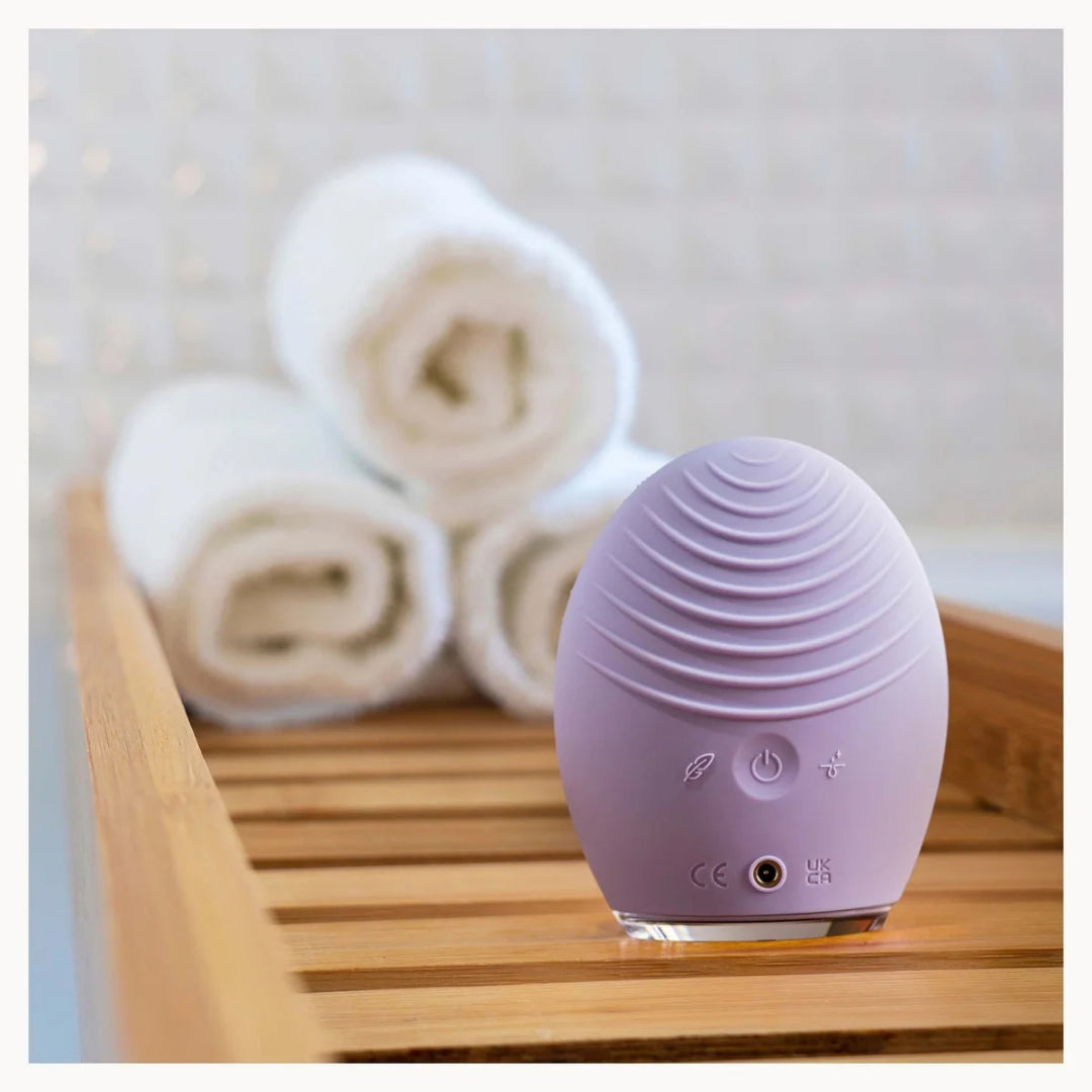 A lavender purple egg-shaped personal massager with a ribbed texture sits on a wooden tray, with rolled white towels blurred in the background against a light tiled wall.