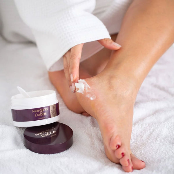 Person applying cream to their foot with a jar of Margaret Debs cream on a white surface.
