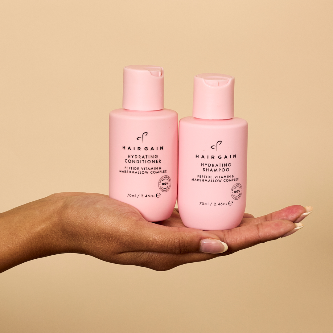 Two pink bottles of Hairgain shampoo and conditioner held in a hand against a beige background