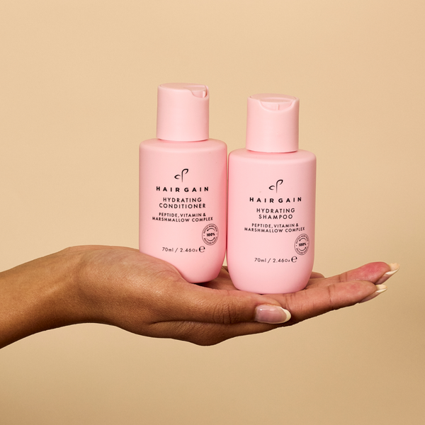 Two pink bottles of Hairgain shampoo and conditioner held in a hand against a beige background