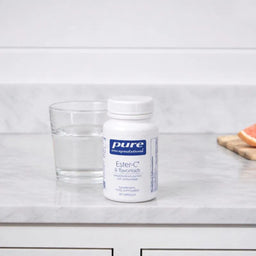 A white bottle of Pure Encapsulations Ester-C & Flavonoids dietary supplement stands on a light marble countertop next to a clear glass of water, with a blurred citrus slice in the background.