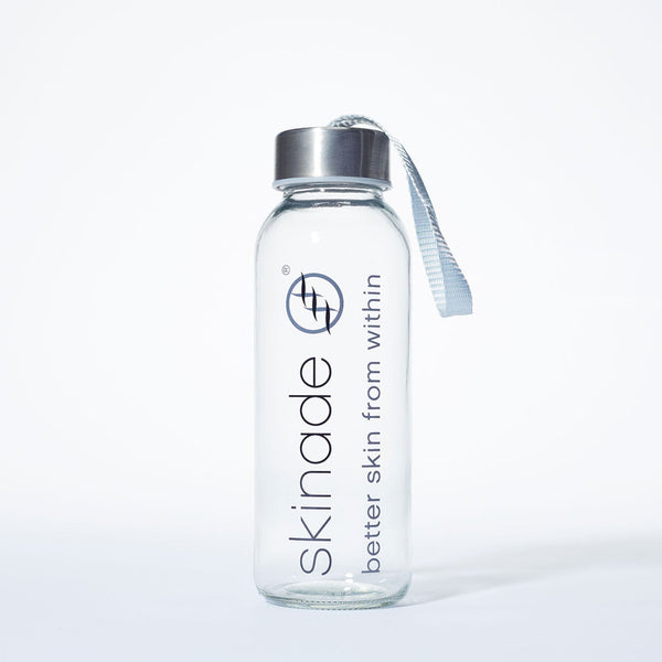 A clear glass water bottle with a silver cap and a light blue-gray carrying strap, featuring the brand name 'skinade' printed vertically in black on the front, set against a clean white background.