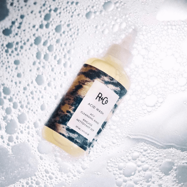 a bottle of R+Co Acid Wash Acv Cleansing Rinse covered in soap suds 